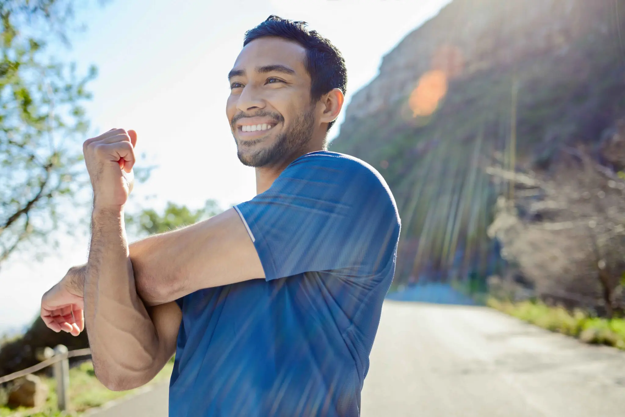 GettyImages-1359149467_11zon (1) A man in a blue shirt stretches his arm and smiles outdoors on a sunny day, with trees and a mountain in the background.