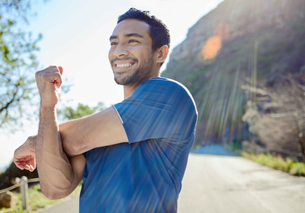 A man in a blue shirt stretches his arm and smiles outdoors on a sunny day, with trees and a mountain in the background.